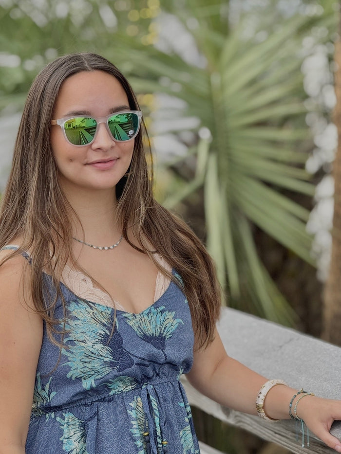 Woman wearing sunglasses and a blue floral dress sitting outdoors with greenery in the background