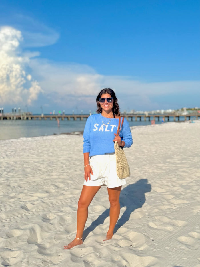 Woman in a blue sweatshirt with 'SALT' on a beach with sunglasses