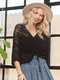 A woman wearing a black crochet knit v-neck sweater with three-quarter length sleeves and a relaxed fit. She has accessorized with a beige hat and has her hair styled in loose waves.