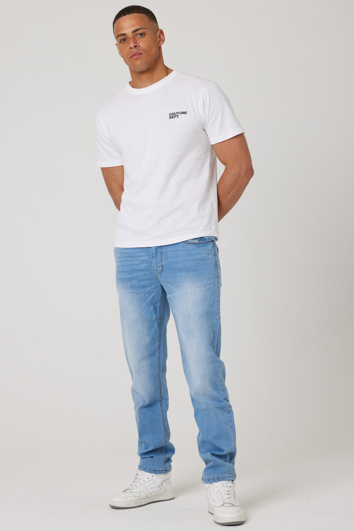A person stands against a plain background, wearing a white T-shirt with small black chest text, Beckenham Straight Fit Stretch Denim Jeans in ice blue, and white sneakers, with hands clasped behind their back.