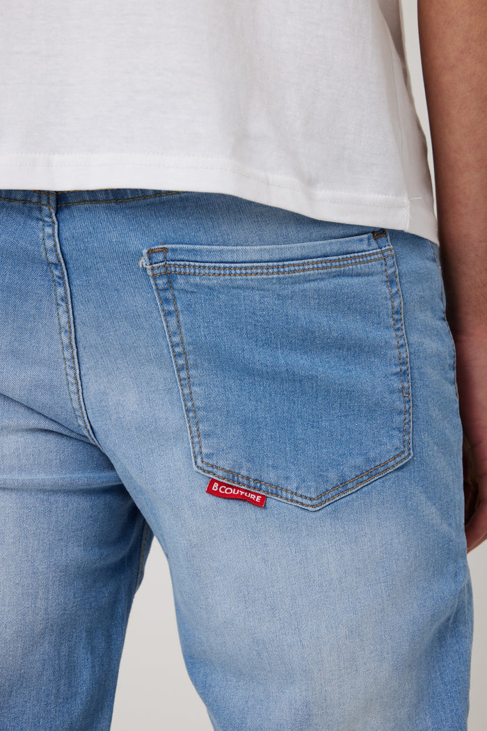 A close-up of someone wearing Beckenham Straight Fit Stretch Denim Jeans in Ice Blue with a red COUTURE label on the back pocket, paired with a white t-shirt.