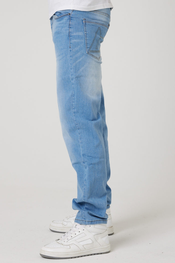 A person wearing Beckenham Straight Fit Stretch Denim Jeans in ice blue and white sneakers stands against a plain white background, shown from the waist down in side profile.