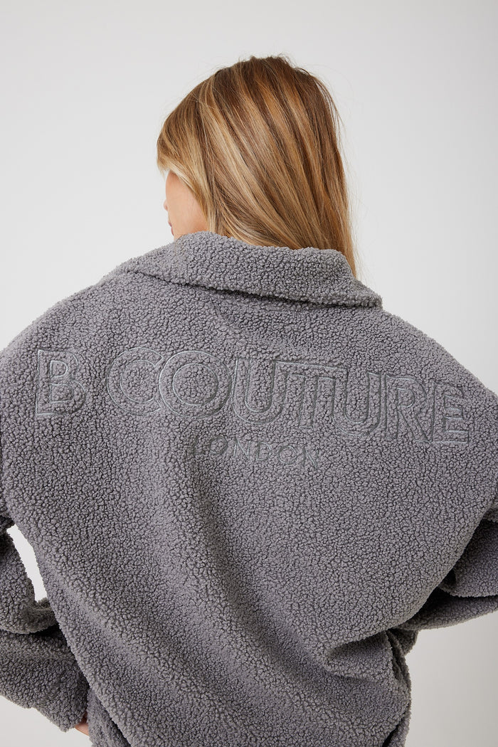 A person with long, light brown hair is shown from the back wearing the grey Belmore Borg Jumper & Leggings Set, featuring a textured teddy fleece and large B. COUTURE embroidery on the back.