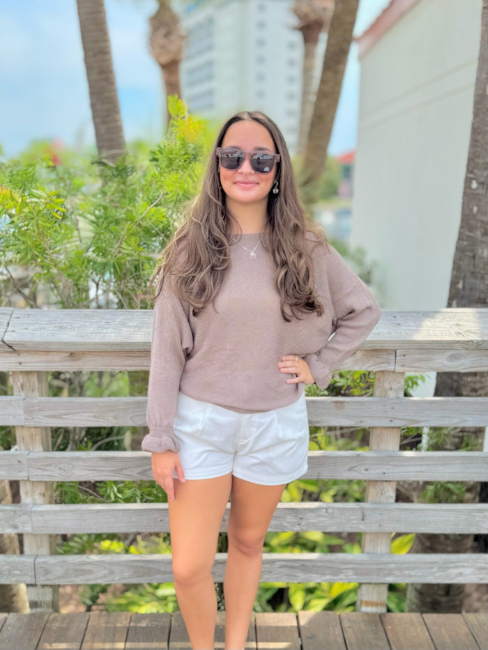 Woman wearing sunglasses and a beige sweater with white shorts standing on a wooden deck with palm trees in the background.