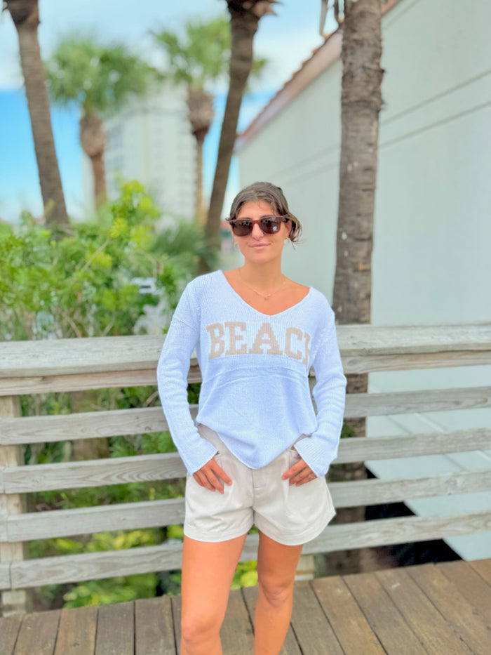 Woman wearing a white 'BEACH' shirt and sunglasses, standing on a wooden deck with palm trees in the background.