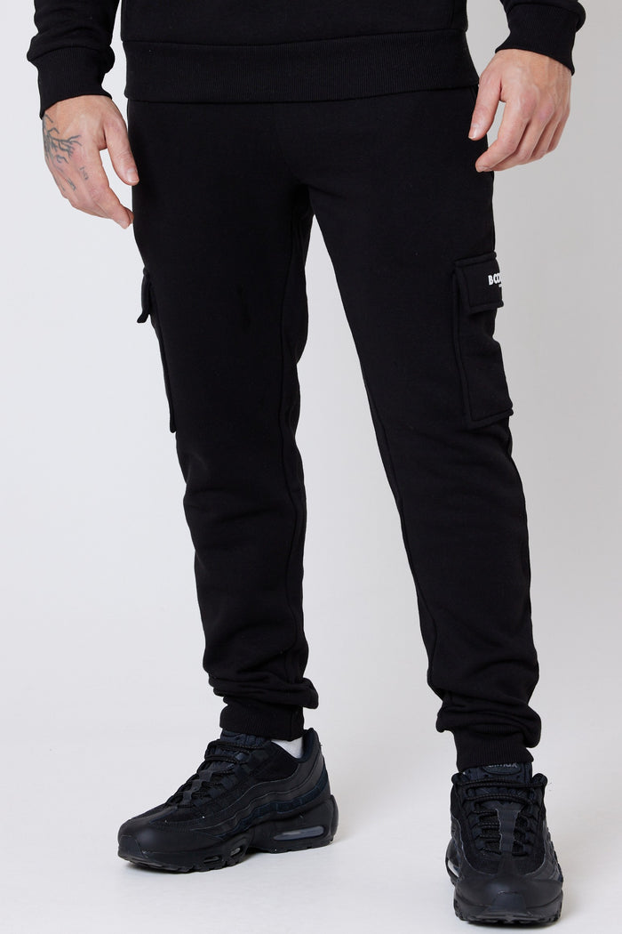 A male model wearing a pair of black fleece cargo joggers with a regular fit, side pockets, and cuffed ankles. He is also wearing a dark-colored top and black sneakers.