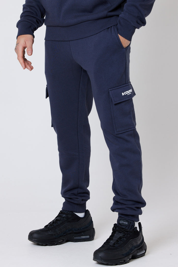 A male model wearing a pair of navy fleece cargo joggers with a regular fit, side pockets, and cuffed ankles. He is also wearing a dark-colored top and black sneakers.