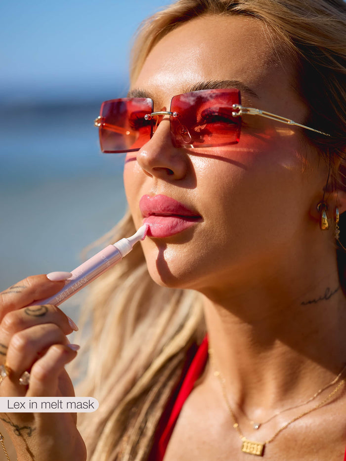A person with long hair applies Tarte Cosmetics Melt Mask to their lips, wearing pink-tinted sunglasses and gold jewelry, set against a blurred outdoor background. Text: Lex in melt mask.