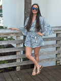 Blue and ivory leaf-print romper with a flattering surplice V-neckline, wide kimono sleeves, an elastic drawstring waist with tassel ties, and relaxed shorts, styled with mirrored blue sunglasses.