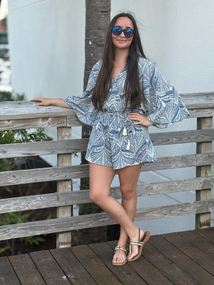 Blue and ivory leaf-print romper with a flattering surplice V-neckline, wide kimono sleeves, an elastic drawstring waist with tassel ties, and relaxed shorts, styled with mirrored blue sunglasses.