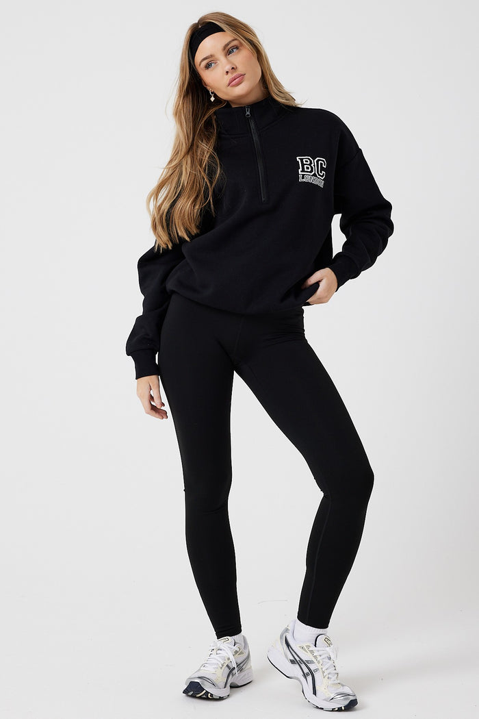 A woman with long hair poses against a plain background wearing the Collingwood Half-Zip Sweatshirt & Leggings Set in black, paired with white sneakers and a black headband.