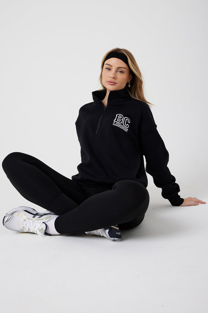 A woman sits relaxed on the floor in the Collingwood Half-Zip Sweatshirt & Leggings Set - Black, paired with white sneakers and a black headband—an effortlessly chic athleisure look against a plain white background.