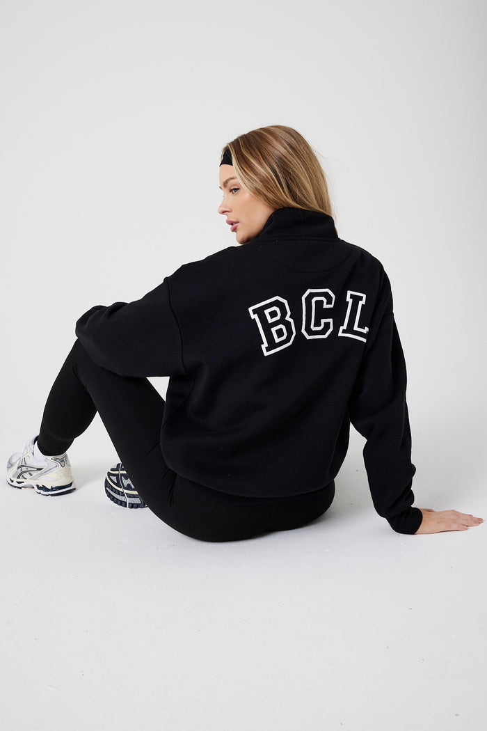 A woman with long hair sits on the floor facing away, wearing the Collingwood Half-Zip Sweatshirt & Leggings Set in black with "BCL" on the back, paired with white sneakers. The background is plain white.