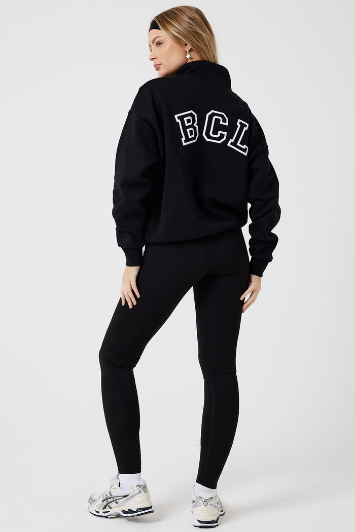A woman showcases the Collingwood Half-Zip Sweatshirt & Leggings Set in black, featuring an oversized sweatshirt with bold white BCL letters on the back and high-waisted leggings, styled with white sneakers against a plain white background.