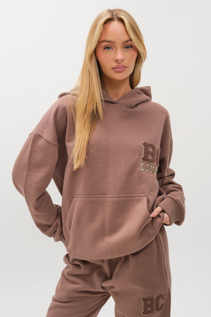 A woman with long blonde hair wears the Etty Fleece Tracksuit in Mink Brown, featuring matching relaxed-fit hoodie and joggers with BC London text patches. She stands against a plain white background, one hand in her pocket.