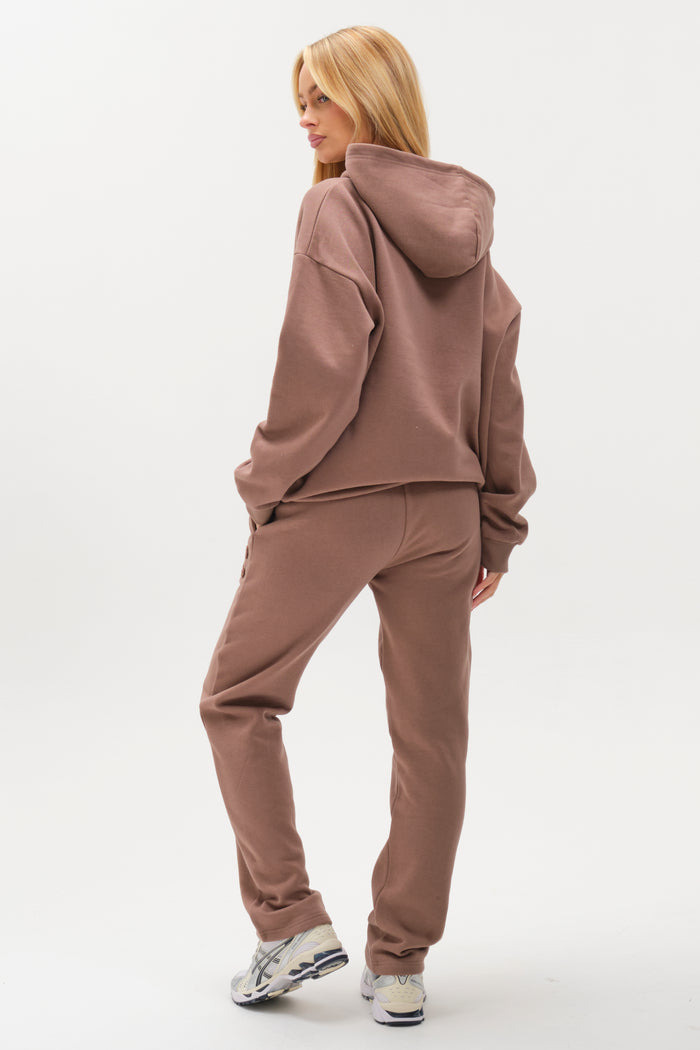 A person with long blonde hair wears the Etty Fleece Tracksuit in Mink Brown, featuring a loose hoodie and matching relaxed fit joggers. They stand slightly turned away on a plain white background, paired with white sneakers for a cozy vibe.