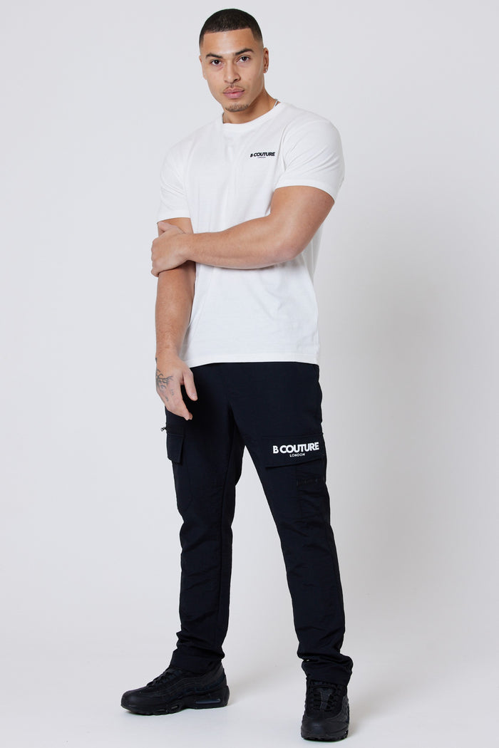 A man stands against a plain white background, wearing a grey Hartfield Regular Cotton T-Shirt from the 3-Pack set (Grey/Stone/Cream) and black cargo pants with "B COUTURE" branding, paired with black sneakers. He looks at the camera neutrally.