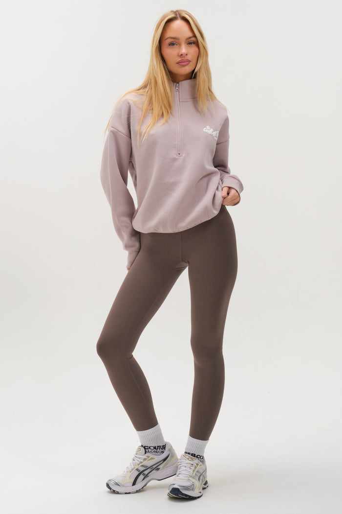 A woman with long blonde hair models the Hatherley Half-Zip Sweatshirt & Leggings Set in Stone, paired with white socks and sneakers. She stands confidently against a plain white background in this relaxed, cosy ensemble.