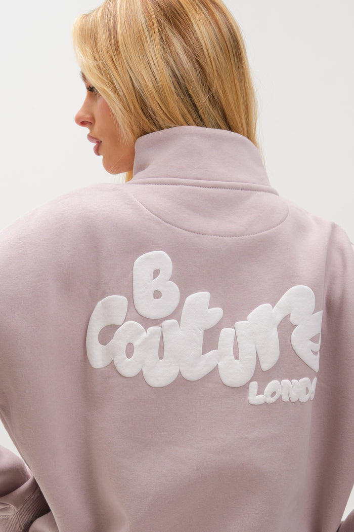 A woman with blonde hair models the Hatherley Half-Zip Sweatshirt & Leggings Set in Stone, turned slightly away to show the relaxed fit and large white "B Couture LONDON" text printed on the back.