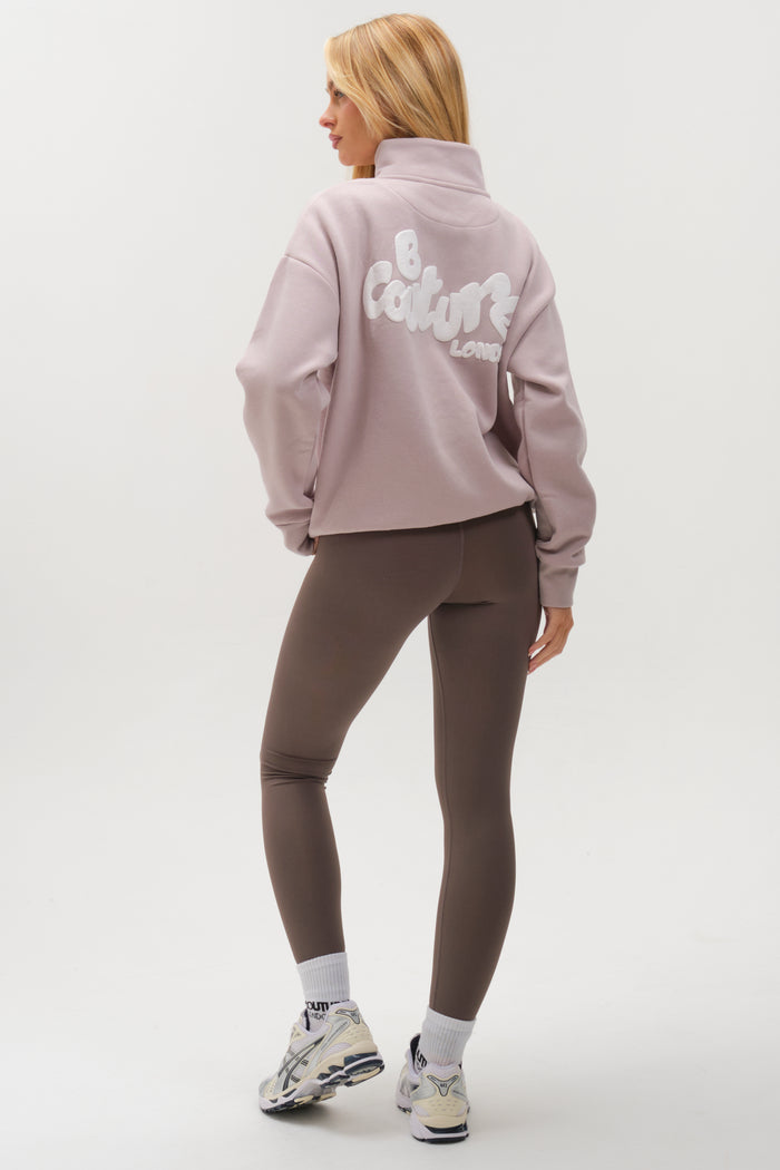 A woman faces away, wearing the Hatherley Half-Zip Sweatshirt & Leggings Set in Stone, paired with white socks and sneakers for a cosy look against a plain white background.