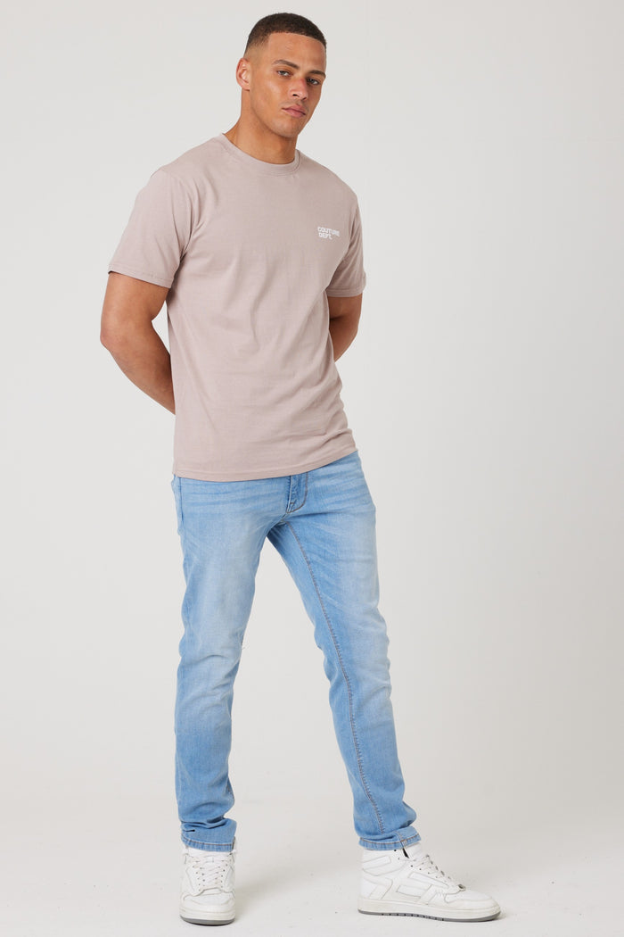 A man stands against a plain white background, wearing the Haydon Walk Regular Fit Cotton T-Shirt in beige, paired with light blue jeans and white sneakers. He clasps his hands behind his back and looks forward with a neutral expression.