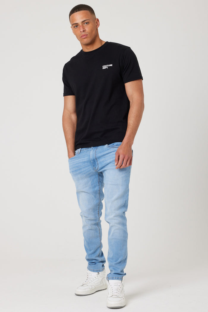 A man stands against a plain white background wearing the Haydon Walk Regular Fit Cotton T-Shirt in black, paired with light blue jeans and white sneakers. His left hand is in his pocket as he looks slightly toward the camera.