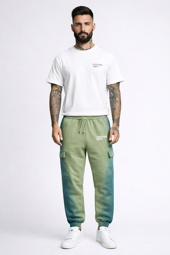 A man with tattoos wears a white T-shirt, Heavy Weight Oversized Couture Department Joggers in green with a tapered leg, and white sneakers, standing against a plain light background.