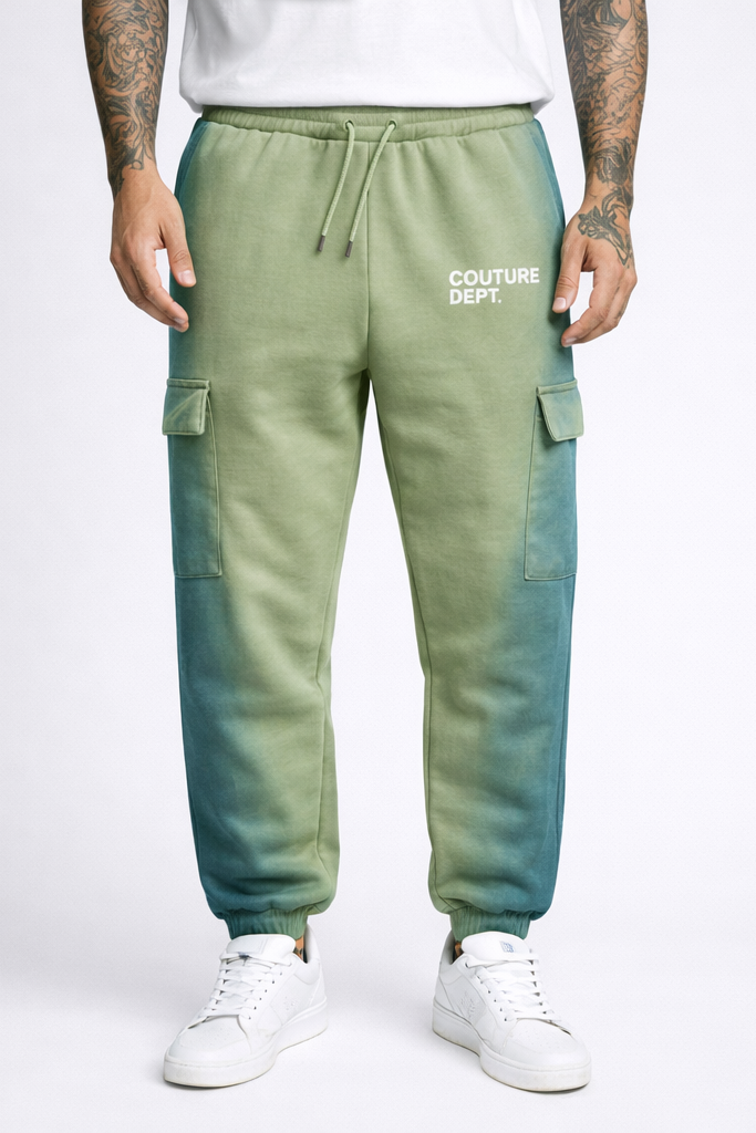 A person wears green Heavy Weight Oversized Couture Department Joggers with white COUTURE DEPT. print on the left thigh, paired with white sneakers and a white shirt. Their arms and hands feature tattoos.