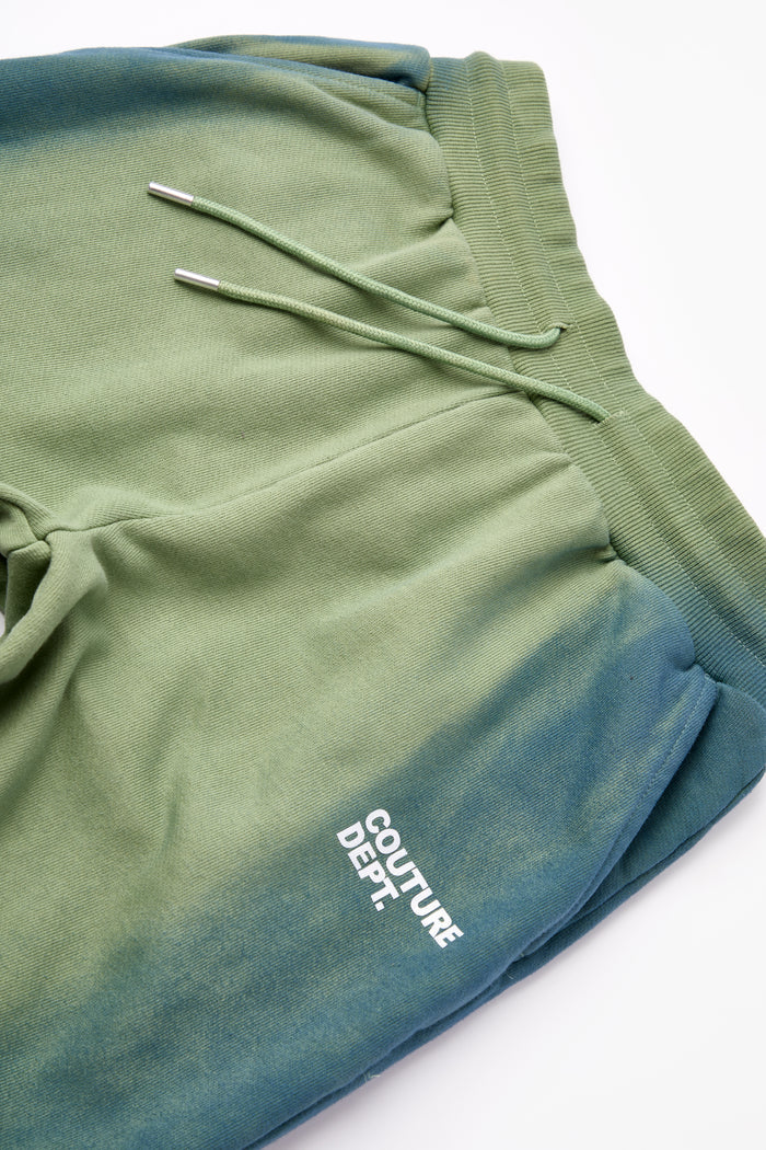 The Heavy Weight Oversized Couture Department Joggers in green feature a blue and green ombré design, elastic waistband with green drawstring, and white Couture Dept. logo on the left thigh. Tapered leg style, shown flat on a white background.