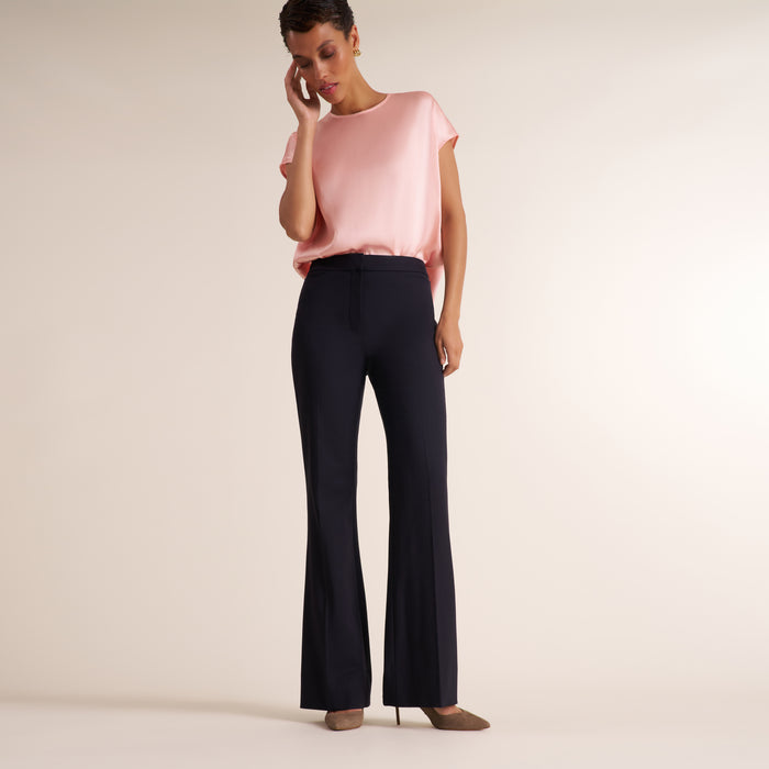 A model wearing a light pink short-sleeve top and dark navy flared trousers, standing against a plain beige studio background. | Lead