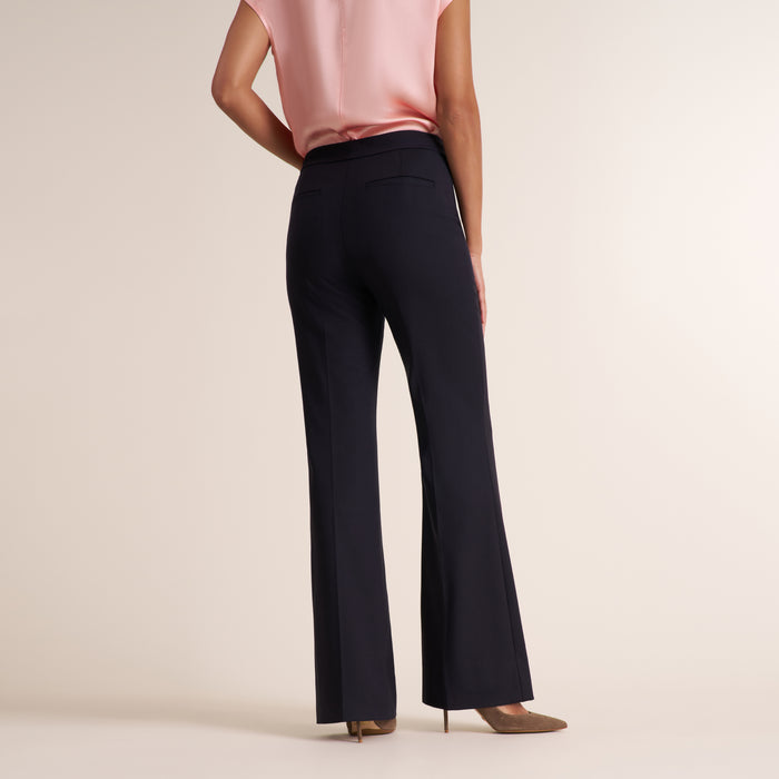 A model from the back wearing high-waisted, wide-leg navy trousers, a light pink satin top, and brown pointed-toe heels against a warm beige studio background.