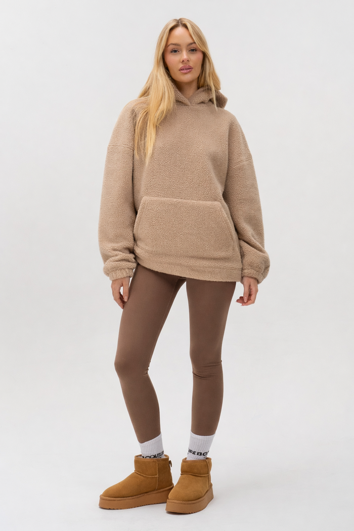 A woman with long blonde hair poses casually against a plain background, wearing the Isla Sherpa Hoodie & Leggings Set in beige and brown, paired with white socks and tan ankle boots for a cosy, relaxed look.