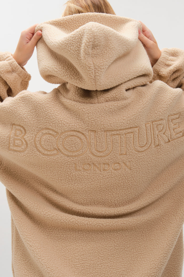 A person faces away, holding up the hood of the beige/brown Isla Sherpa Hoodie & Leggings Set for cozy warmth, showing off its oversized, fuzzy sherpa style.