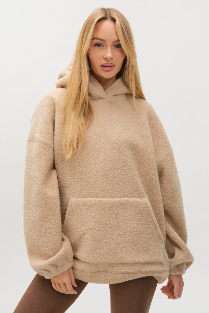 A woman with long blonde hair models the Isla Sherpa Hoodie & Leggings Set in beige/brown, featuring an oversized fit, front pocket, and hood for cozy warmth, posed against a plain light background.