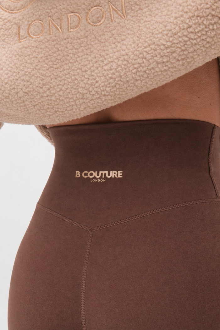 Close-up of a person in the Isla Sherpa Hoodie & Leggings Set - Beige/Brown, featuring high-waisted brown leggings with B COUTURE LONDON on the waistband and a cosy fleece hoodie embroidered with LONDON, draped over their shoulders.