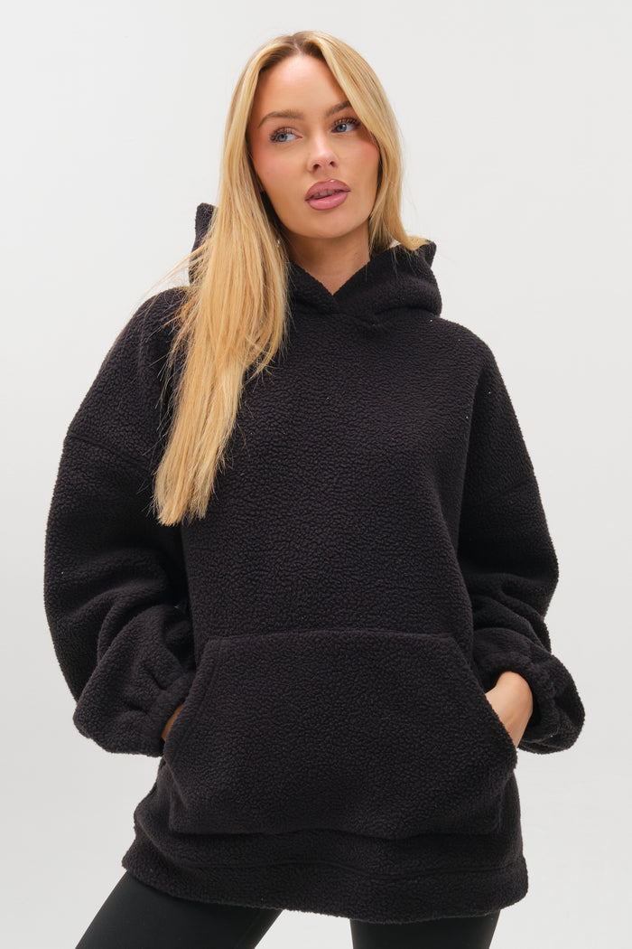 A woman with long blonde hair wears the Isla Sherpa Hoodie & Leggings Set in black, featuring a plush fleece oversized hoodie with a front pocket, standing against a plain white background, radiating cosy warmth.
