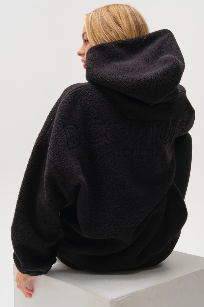 A blonde woman sits on a white block, facing away, wearing the Isla Sherpa Hoodie & Leggings Set - Black with "B COUTURE LONDON" embroidered on the back. The textured set exudes cosy warmth against a plain white background.