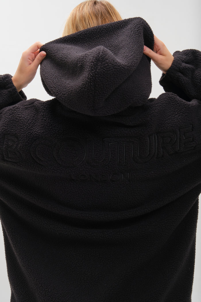 A blonde-haired person faces away, wearing the Isla Sherpa Hoodie & Leggings Set - Black, with B COUTURE LONDON embossed on the back, and holding up the hood with both hands.
