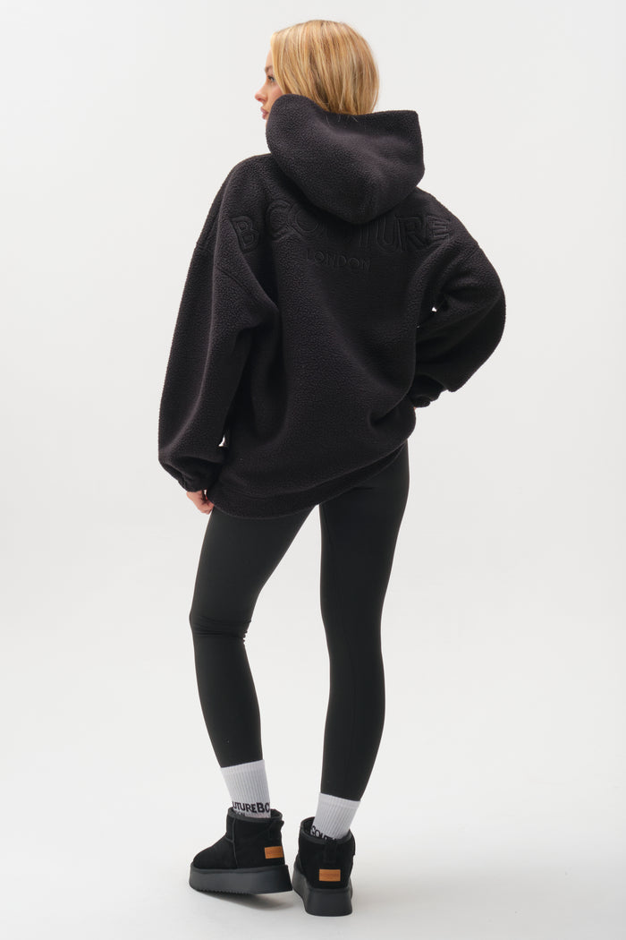 A person with long blonde hair faces away in the Isla Sherpa Hoodie & Leggings Set - Black, styled with white socks and black platform sneakers against a plain white background.