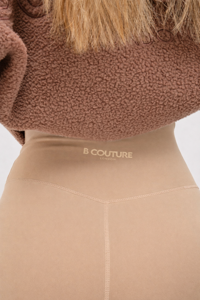 A close-up of someone wearing the Isla Sherpa Hoodie & Leggings Set in brown/beige, featuring high-waisted leggings with B COUTURE LONDON on the waistband, paired with a cosy sherpa oversized hoodie and a brown textured sweater.