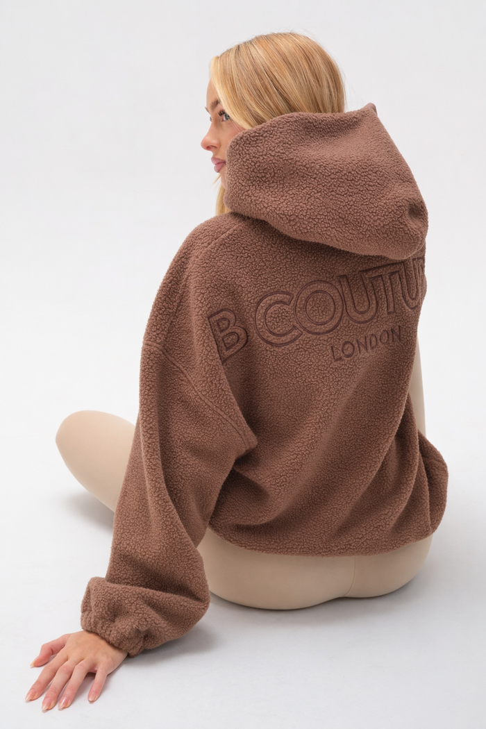 A person with long blonde hair sits on the floor, back turned, wearing the Isla Sherpa Hoodie & Leggings Set in brown/beige, against a plain light background. The hoodie is oversized and features B COUTURE LONDON embroidery on the back.