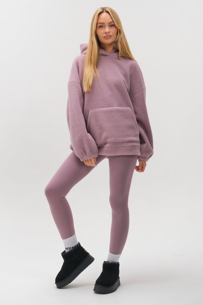 A woman with long blonde hair poses confidently against a white background in the Isla Sherpa Hoodie & Leggings Set - Mauve, paired with black ankle boots and white socks.