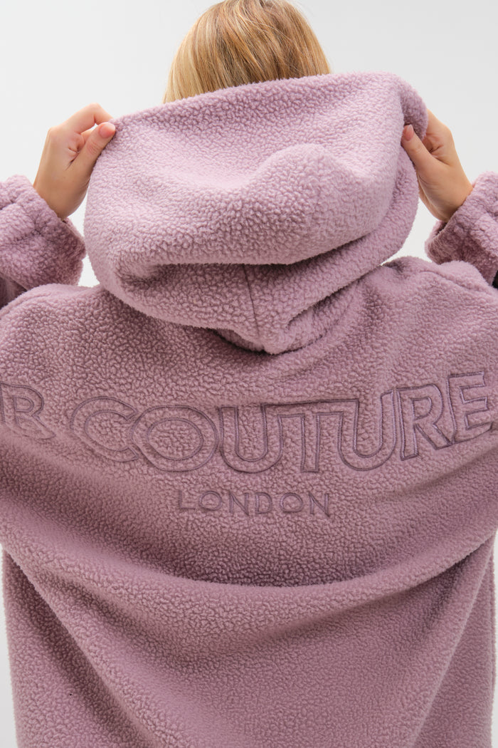 A person lifts the hood of the Isla Sherpa Hoodie in mauve, showing its cosy, textured fleece. Large "COUTURE LONDON" embroidery stands out on the back, while only their hands and hair are visible, highlighting this warm statement set.