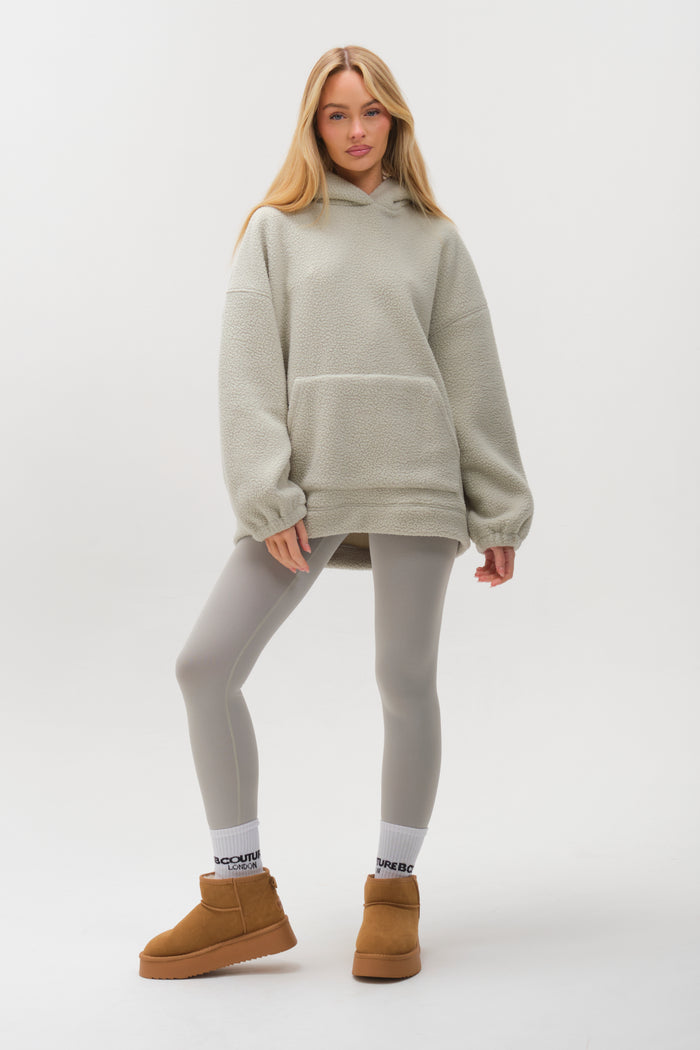 A woman exudes cosy warmth against a plain white background in the Isla Sherpa Hoodie & Leggings Set in teal, paired with white socks and tan boots.