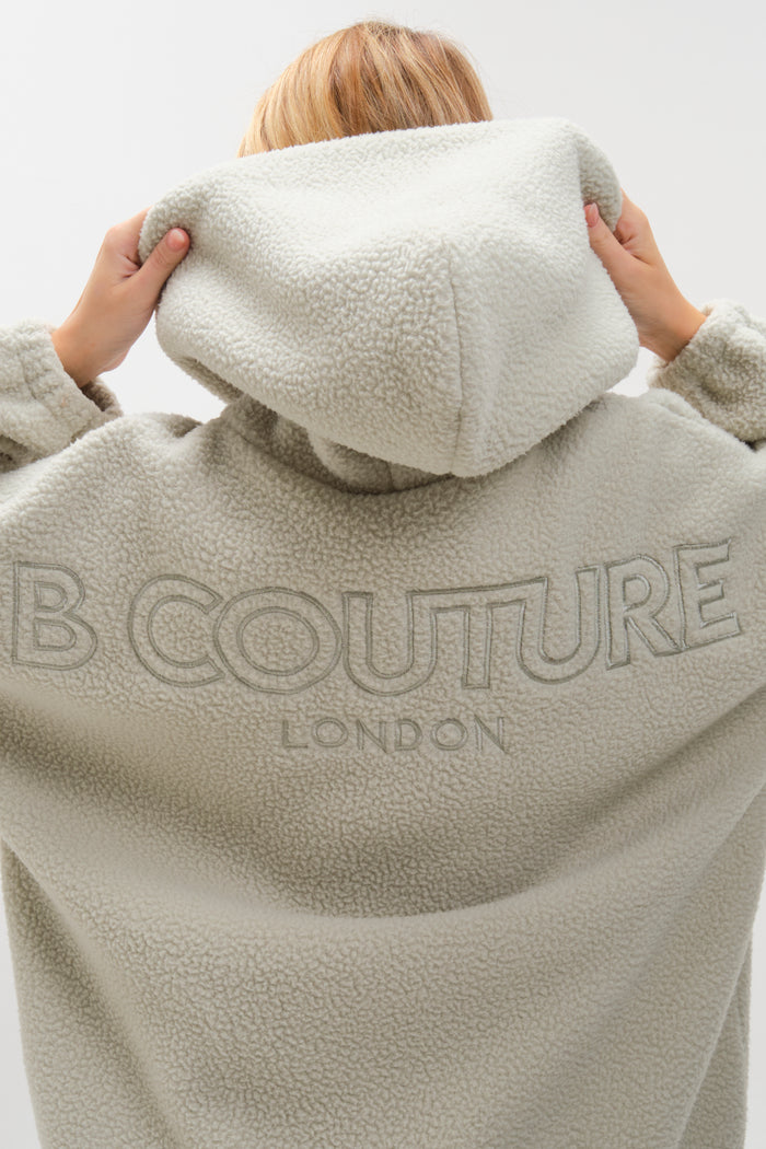 A person faces away, pulling up the hood of the plush Isla Sherpa Hoodie in Teal. "COUTURE LONDON" is embroidered on the back. The plain white background highlights this cosy hoodie and leggings set.