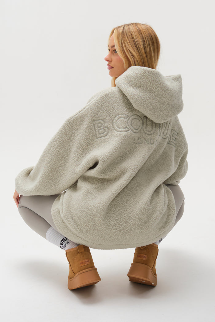 A woman with long blonde hair wears the Isla Sherpa Hoodie & Leggings Set in teal, squatting to show off the B COUTURE LONDON text on the back of her cosy hoodie.