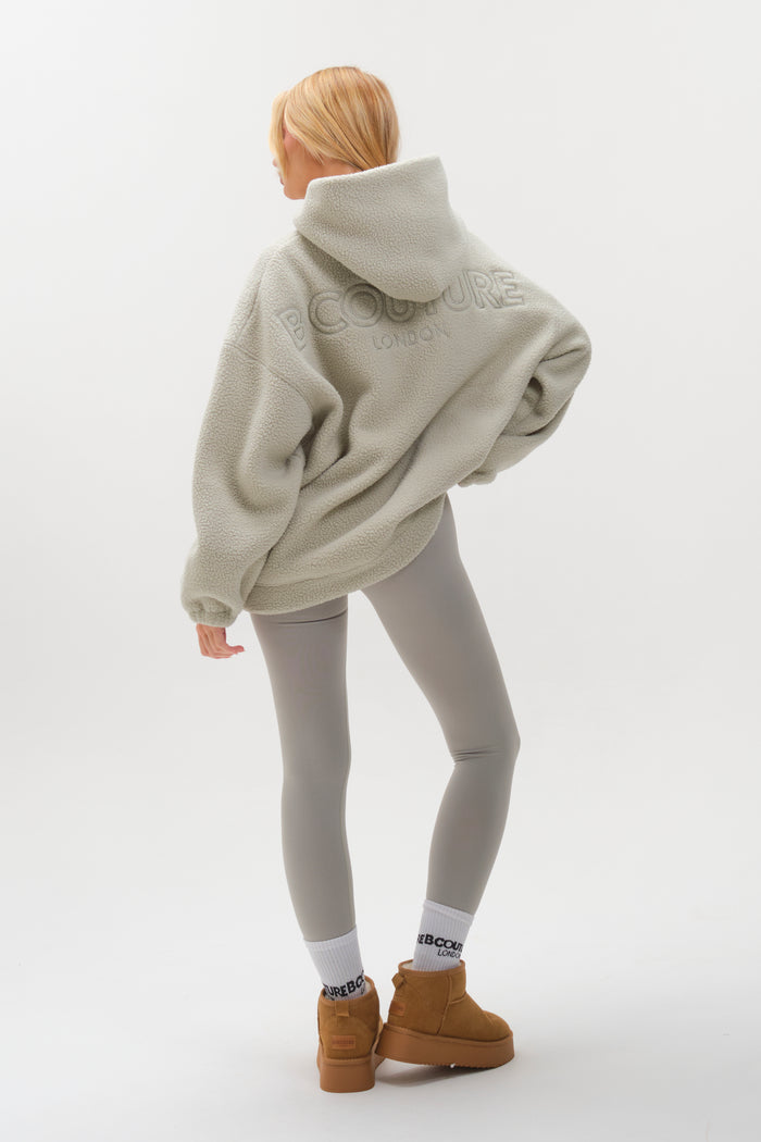 A person with blonde hair wears the Isla Sherpa Hoodie & Leggings Set in teal, paired with white socks featuring black text and tan boots. They stand hand-on-hip, facing away against a plain white background.