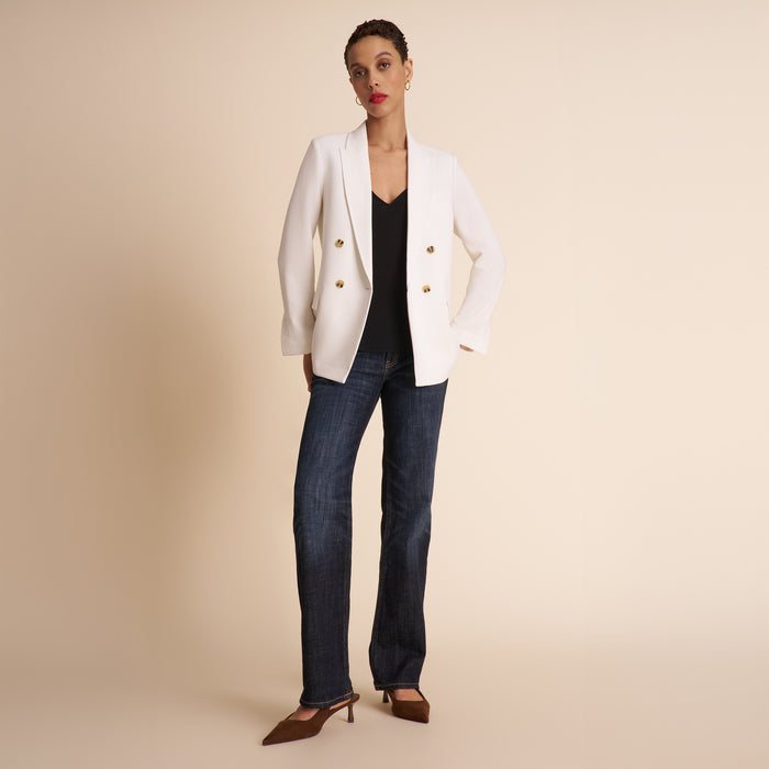 Woman wearing a white blazer, black top, and blue jeans on a beige background | Exclude