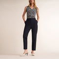 A woman models a sleeveless V-neck top with a black and white floral pattern, paired with dark navy cropped trousers and white slingback heels, against a solid light beige studio background. | Lead