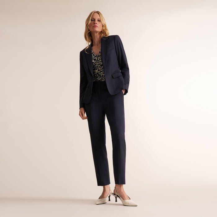 A woman models a dark navy blue two-piece pantsuit, consisting of a single-breasted blazer and matching trousers, paired with a patterned top and white pointed-toe heels, set against a plain beige studio background.